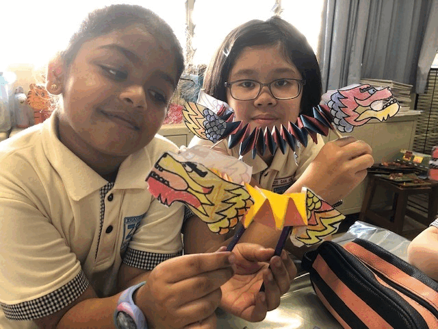 Two children in school uniforms hold colorful paper dragon crafts in a classroom.
